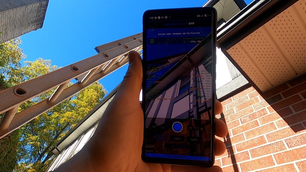 A hand holding a phone running the Verified Reality app, with the ladder and brick wall of the job behind it.